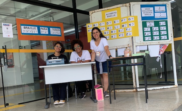 Fotografia de Anna, Samantha e Fernanda, responsveis pela interveno, posam para a foto em frente a cartazes com textos impressos em tinta e em braille.