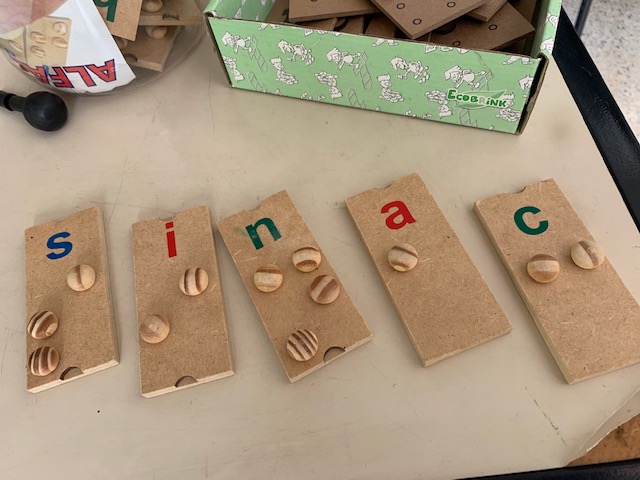 Fotografia de placas de madeira que compem a sigla SINAC, com texto em tinta e com bolinhas que representam letras em braille.