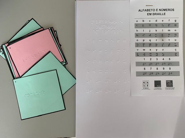 Fotografia de papis coloridos com impresses em braille ao lado de um alfabeto braille, impresso tambm em tinta.